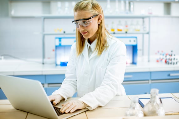 cute-female-researcher-white-lab-coat-protective-goggles-using-laptop-while-working-laboratory